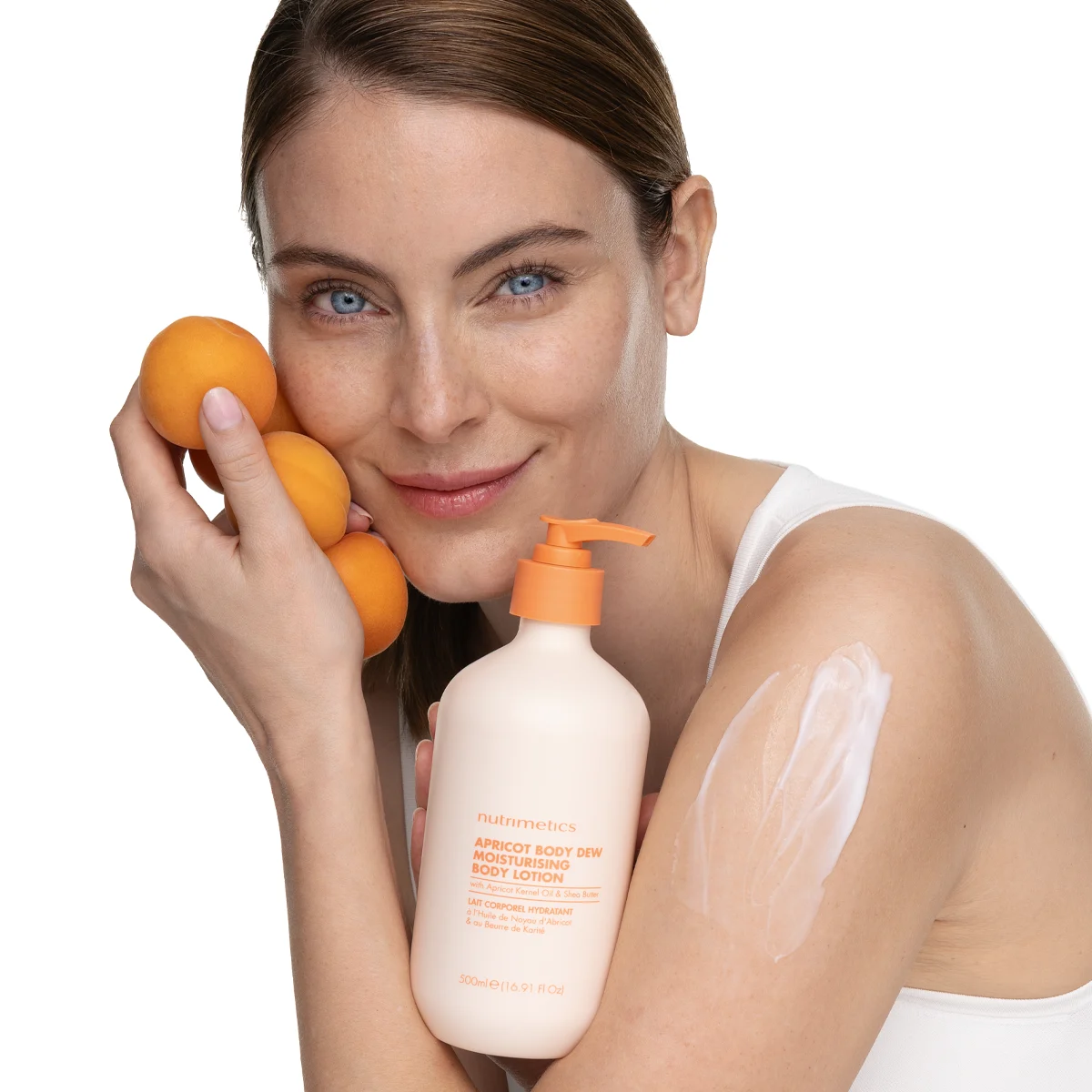 A woman holding fresh apricots next to her face while showcasing Nutrimetics Apricot Body Dew Moisturising Body Lotion on her arm.